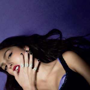olivia rodrigo laying on a purple background, with the tip of her thumb in her mouth. she is wearing rings that spell out "GUTS," with each letter on a different finger.